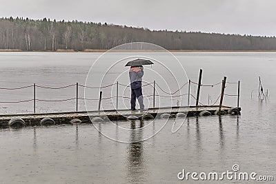 Man talking on phone and walking under umbrella on wooden pier on rainy gray day over of backdrop of lake and forest. Editorial Stock Photo