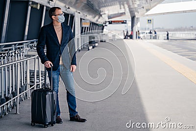 Man traveler poses with suitcase on wheels wears protective mask during pandemic outbreak afraids of dangerous influenza Stock Photo