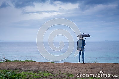 A man with an umbrella in the rain stands on a cliff and looks at the ocean back view. tourist place. Stock Photo