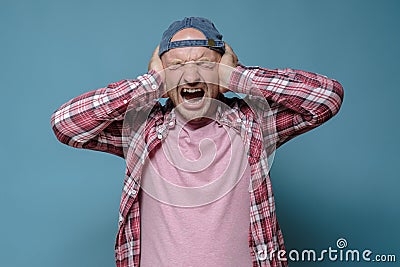 Man is under stress, he is tired of the noise, covers ears with hands and screams nervously. Blue background. Stock Photo