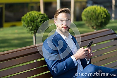 Man use smartphone in park. Manager with mobile phone on sunny outdoor. Businessman in blue suit sit on bench. Modern life and bus Stock Photo