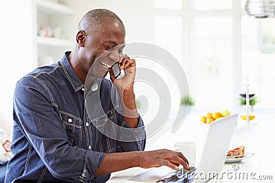 Man Using Laptop And Talking On Phone In Kitchen At Home Stock Photo