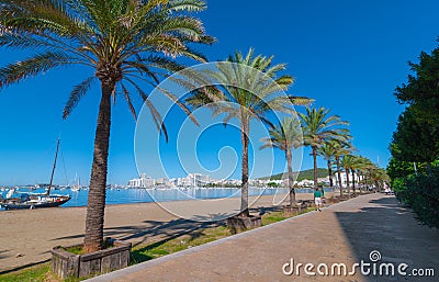 Man walks by an abandoned sailboat on the beach. Rows of palm trees line water`s edge in Ibiza. Stock Photo