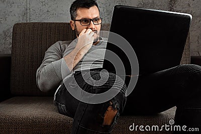 A man watches an adult video on a laptop while sitting on the couch. The concept of porn, men`s needs, pervert, lust, desire, Stock Photo