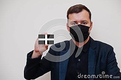 Man wear black formal and protect face mask, hold Cornwall flag card isolated on white background. United Kingdom counties of Stock Photo