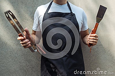 A man wearing black chef`s apron, holding barbecue tools: bbq tongs, spatula Stock Photo