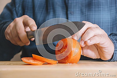 Man wearing a blue shirt slicing a tomato on a wooden board. Cooking concept Stock Photo