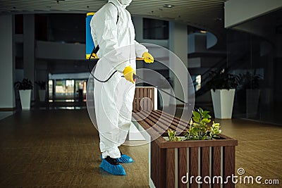 A man wearing protective suit is disinfecting a bench in an empty shopping mall with sanitizing spray. Cleaning up the public Stock Photo