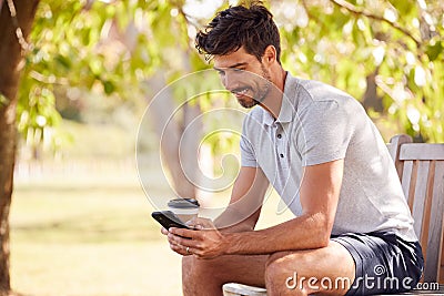 Man Wearing Summer Shorts Sitting On Park Bench Under Tree With Takeaway Coffee Using Mobile Phone Stock Photo
