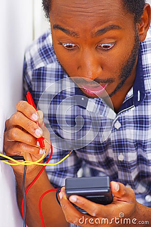 Man wearing white and blue shirt working on electrical wall socket wires using multimeter, electrician concept Stock Photo
