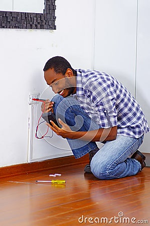 Man wearing white and blue shirt working on electrical wall socket wires using multimeter, electrician concept Stock Photo