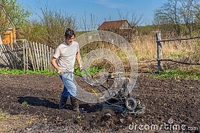 Man in wellingtons with cultivator ploughing ground in sunny day. Farmer plowing kitchen-garden in suburb. Land cultivation, soil Stock Photo