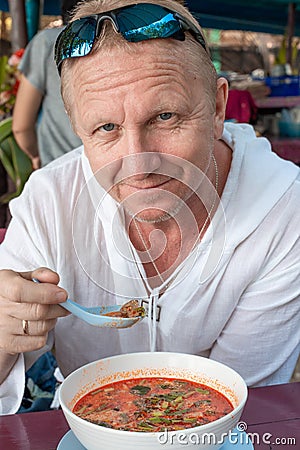 Man in a white tunic eats a spicy Thai soup Tom Yam in a cafe. Gastronomic tourism in Asia. Stock Photo