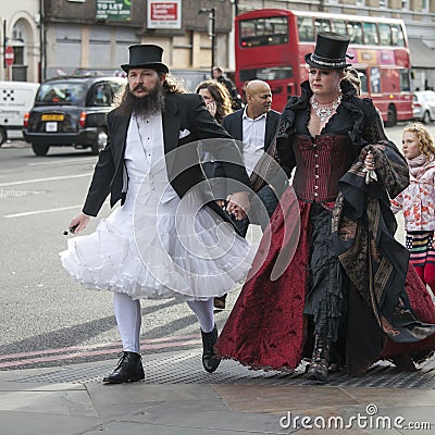 A man in a white wedding dress and a top hat with a woman in a gothic red velvet dress walking down the street in London Bridge Editorial Stock Photo