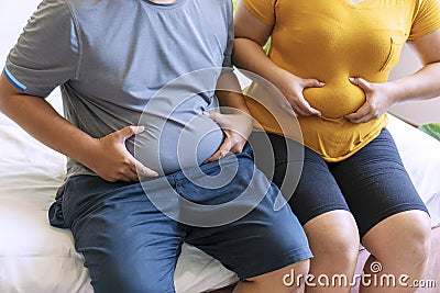 Man and woman holding their big belly while sitting on the bed suffering from extra weight. Couple heavy body size worry Stock Photo