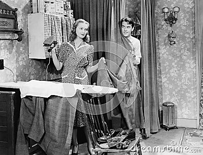 Man and woman standing in a kitchen while she is ironing his pants and he is behind a curtain Stock Photo