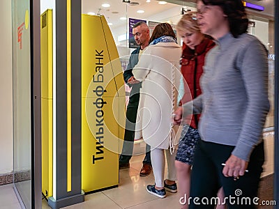 A man and a woman are standing near the yellow terminal with the Tinkoff Bank logo. ATM at the entrance to the mall. Visitors pass Editorial Stock Photo