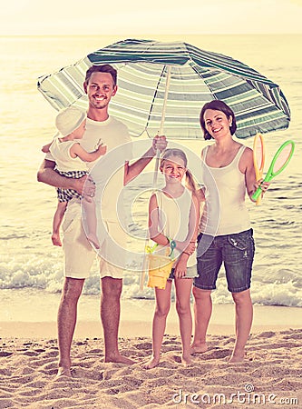 man and woman with two kids standing together under beach umbrel Stock Photo