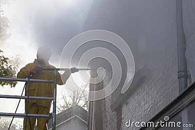 Man in yellow rain suit cleans paint from brick wall of house fa Stock Photo