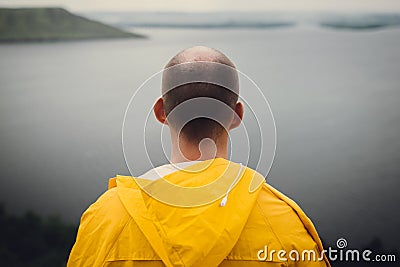 Man in yellow raincoat traveling on cliff and looking at lake in rainy windy day. Atmospheric moment. Wanderlust and travel Stock Photo