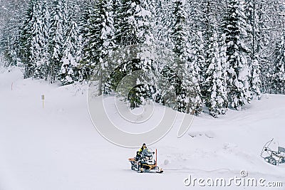 Man in a yellow ski suit rides a snowmobile through a snowy forest at the foot of a hill Editorial Stock Photo