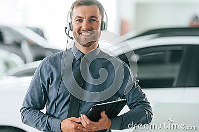 Manager is in the headphones and with notepad. Handsome car dealership worker is with electric vehicle Stock Photo