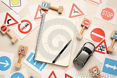Many different road signs, notebook with sketch of roundabout and toy car on white wooden background, flat lay. Driving school Stock Photo
