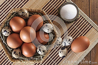 Many eggs. Large brown chicken eggs and small motley quail eggs on straw in wicker basket and salt in salt shaker on wooden table Stock Photo