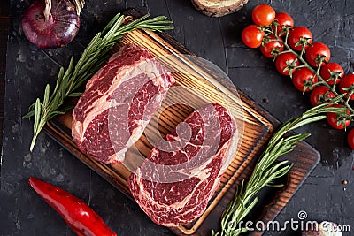 Marble ribeye steak on a cutting board with rosemary, peas, onions, garlic, red tomatoes on a branch on a concrete dark background Stock Photo