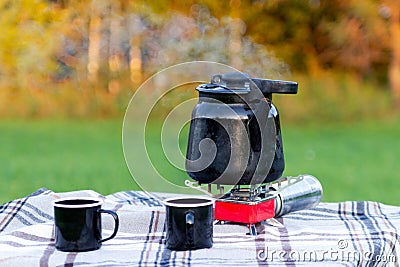 Marching hot iron black smoking teapot on red gas burner and iron mugs are standing on the table on white checkered tablecloth Stock Photo