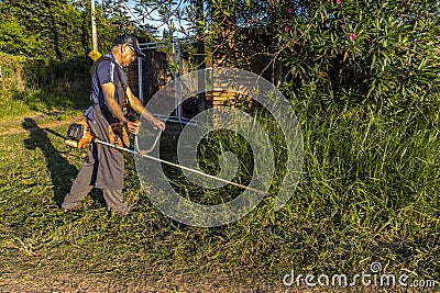 Worker, with a manual gasoline brush cutter, cuts the weeds that grew with the rains in front of the gate Editorial Stock Photo