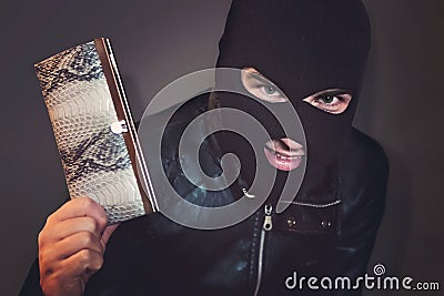 Masked criminal at the black background grins looking at the viewer and showing a purse in hand. Evil-doer is glad to Stock Photo