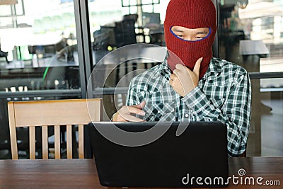 Masked hacker wearing a balaclava looking a laptop and stealing important information data. Network security and privacy crime con Stock Photo