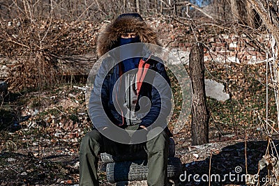 A masked man, hooded jacket, and gloved military pants sits and looks into the camera Stock Photo