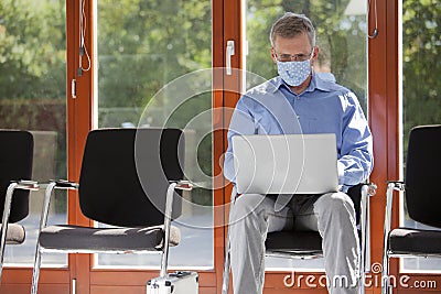 Mature businessman with face mask working with laptop in an empty waiting room of an office or hospital Stock Photo