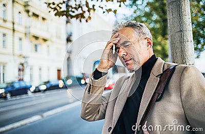 Mature businessman in pain standing on a street in city, holding forehead. Stock Photo