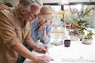 Mature Couple Reviewing And Signing Domestic Finances And Investment Paperwork In Kitchen At Home Stock Photo