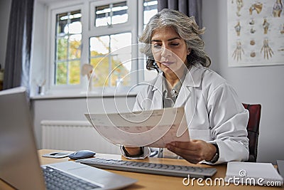 Mature Female GP Wearing White Coat At Desk In Doctors Office Look At ECG Readout Stock Photo