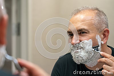Mature gray-haired man restyling his beard himself at home using razor Stock Photo