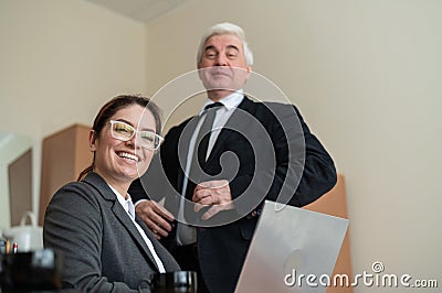 A mature male boss praises a subordinate. A woman in a suit works at a laptop at a desk. Friendly colleagues chat in the Stock Photo