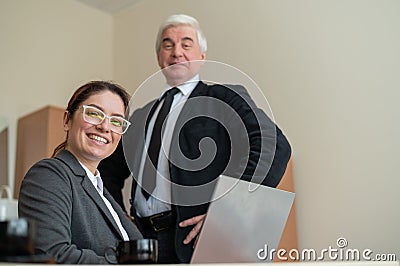 A mature male boss praises a subordinate. A woman in a suit works at a laptop at a desk. Friendly colleagues chat in the Stock Photo