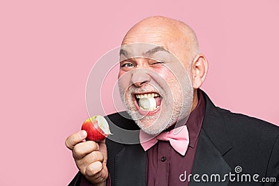 Mature man granddad in suit eat apple on pink background. Portrait of a funny elderly man eating an apple. Stock Photo