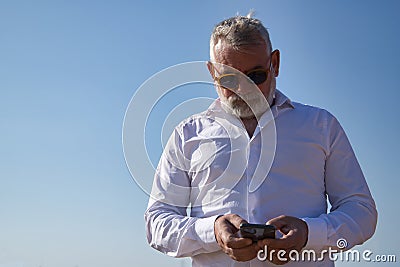Mature man, gray haired, bearded, sunglasses, white shirt, checking social networks on his cell phone, outdoors. Concept Stock Photo