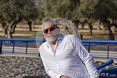 Mature man, gray-haired, bearded, sunglasses, white shirt, leaning on a railing outdoors. Concept grandfather, sugar daddy, Stock Photo
