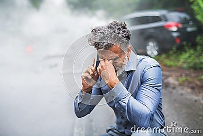 Mature man making a phone call after a car accident, smoke in the background. Stock Photo