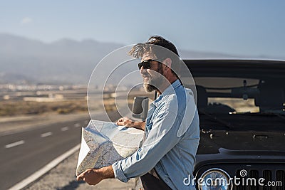 Mature man in sunglasses searching direction on location map while standing beside bonnet of jeep at highway. Handsome man reading Stock Photo
