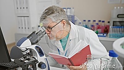 Mature scientist using microscope in laboratory beside computer while holding notebook Stock Photo