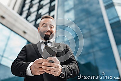 Mature successful entrepreneur using smartphone, standing outdoors against modern business center, low-angle shot Stock Photo