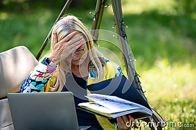 Senior woman with laptop and documents working in garden on rocking couch, green home office concept Stock Photo