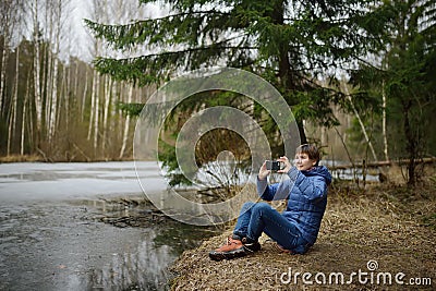 Mature woman is resting on shore of forest lake on early spring. Person takes pictures of nature with smartphone. Outdoor activity Stock Photo